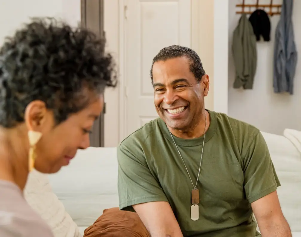 Talking together in the living room of their home, the mature adult veteran smiles as his mature adult wife looks down.
