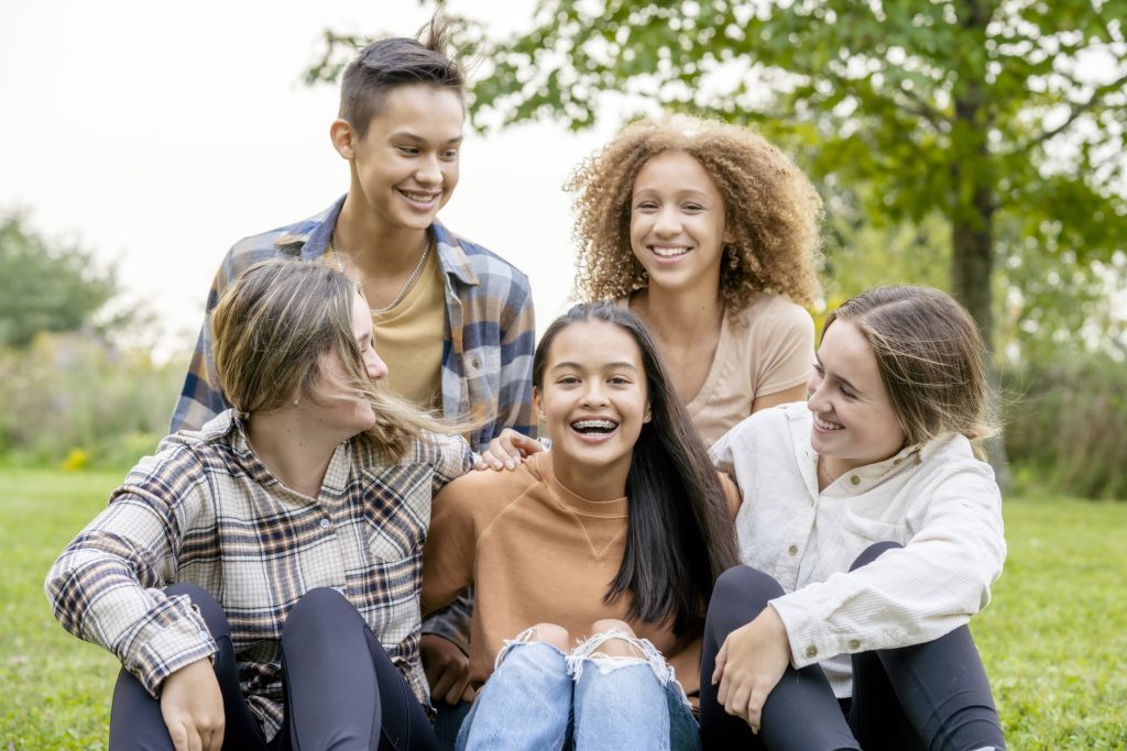 A small group of ethnically diverse high school students sit together outside while on break. They are huddled in closely with their arms around one another, each dressed casually and laughing with each other.